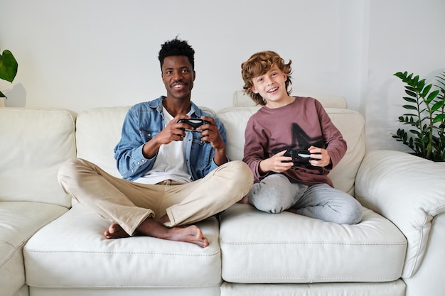Full body smiling multiracial male and teenage boy in casual clothes playing video game with joysticks while sitting with legs crossed on soft couch