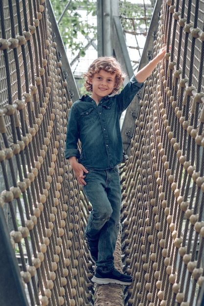 Full body of smiling preteen boy walking on wooden log and holding ropes and looking at camera while passing track in rope adventure park