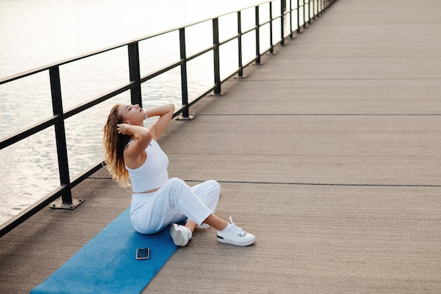 Full body of smiling relaxed female sitting on yoga mat on bridge near river