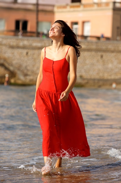 Full body smiling woman in red dress walking in sea barefoot