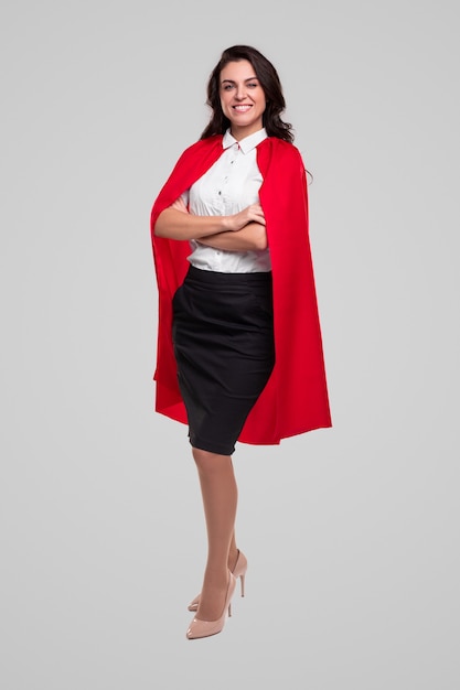Full body of successful stylish businesswoman in elegant office outfit and red superhero cloak looking at camera and smiling against gray background