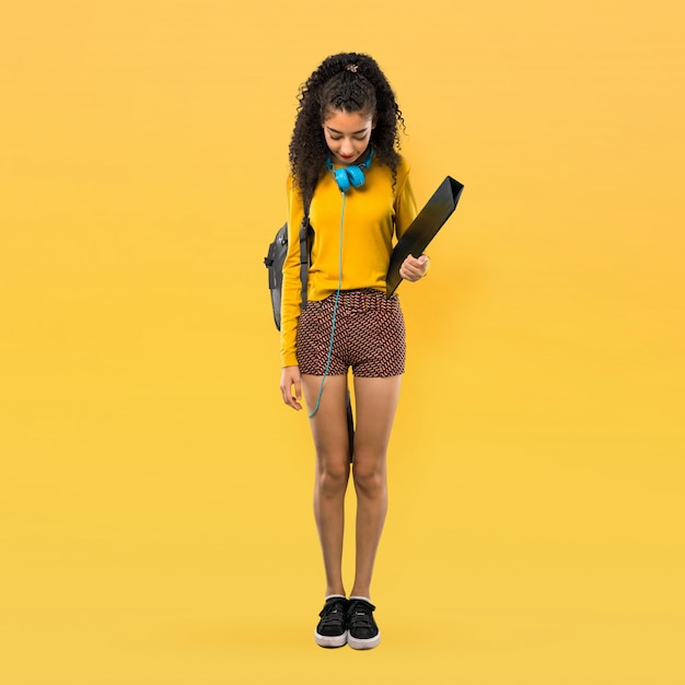 Full body of Teenager student girl with curly hair standing and looking down on yellow background