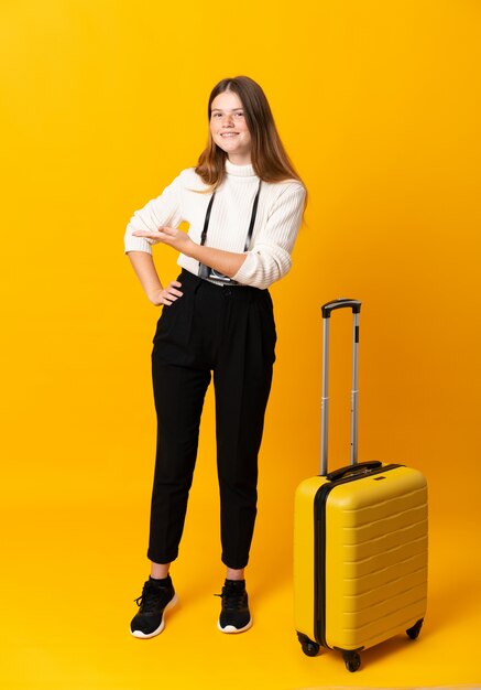 Full body of traveler teenager girl with suitcase over isolated yellow extending hands to the side for inviting to come
