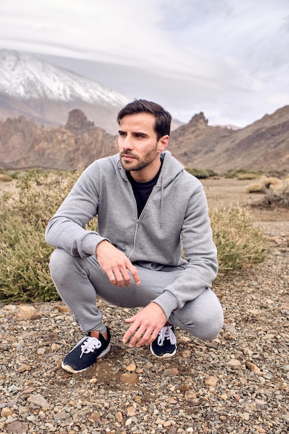 Full body of unshaven pensive male in activewear and sneakers sitting on squat in rocky terrain near Teide volcano located in Tenerife of Canary Islands in Spain and looking away