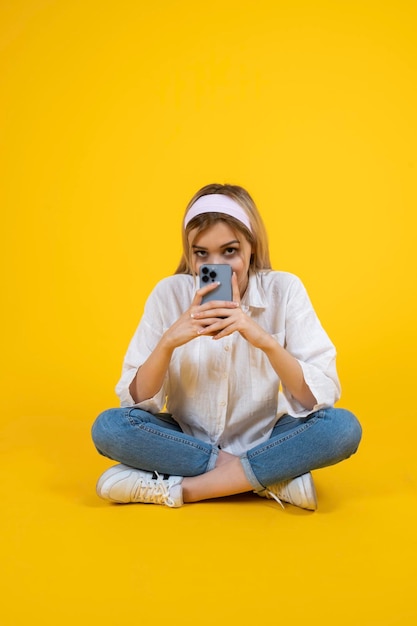 Full body view young blonde girl using smartphone sitting on floor crosslegged copy space