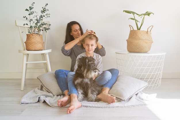 Full body of woman doing hairstyle to cute son while sitting on flow with obedient Miniature Schnauzer dog at home