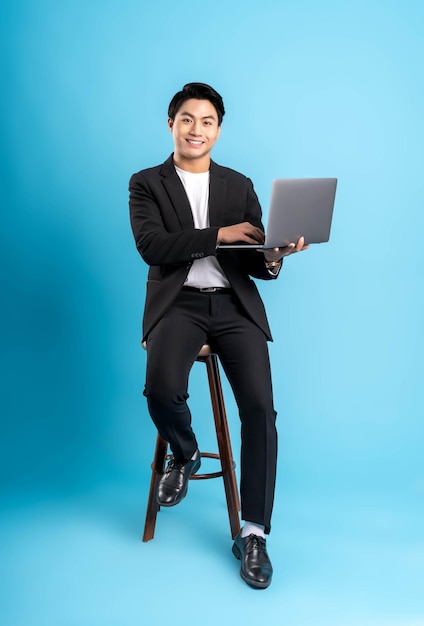 Молодой бизнесмен в жилете, использующий ноутбук и позирующий на синем фоне