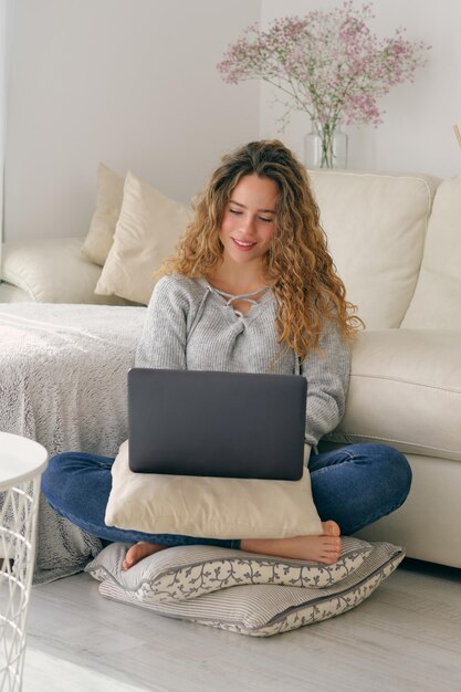 Full body of young female freelancer in casual clothes sitting on floor and working remotely on netbook in living room