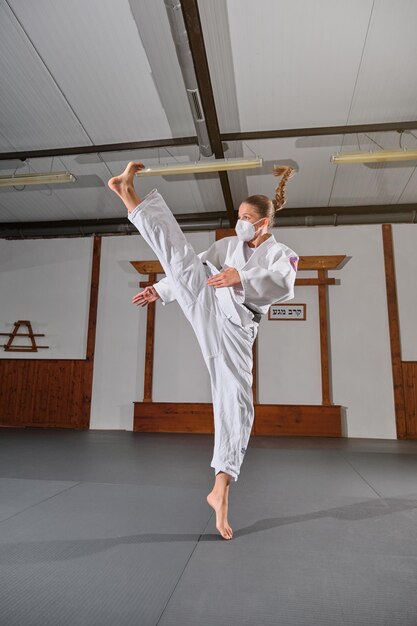 Full body of young female in kimono and medical mask practicing Krav Maga in gym room