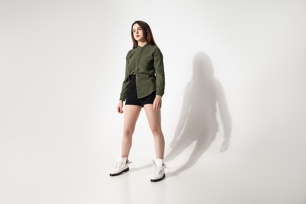 Full body young female in trendy shirt and shorts looking away while standing against gray background