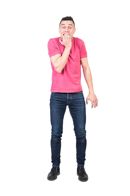 Full body of young nervous male with dark hair in t shirt and jeans biting nails and looking at camera against white background