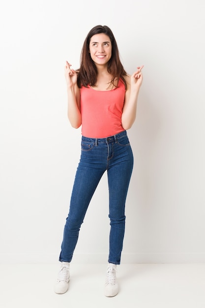 Full body young woman on white wall crossing fingers for having luck