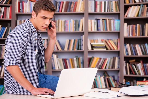 Полная концентрация на учебе. Уверенный молодой человек, работающий на ноутбуке и разговаривающий по мобильному телефону, опираясь на стол и в шрифт книжной полки