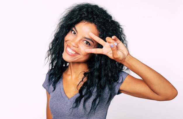 Портрет обрадованной африканской этнической женщины в анфас с миндалевидными темными глазами и густыми волосами, смотрящую в камеру и показывающую v-образный знак