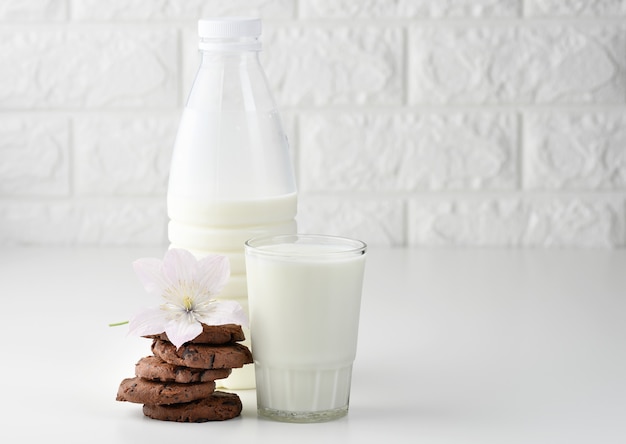 Una tazza piena di latte e una bottiglia di plastica trasparente con latte, accanto a una pila di biscotti rotondi con scaglie di cioccolato su un tavolo bianco