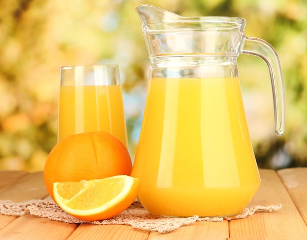 Full glass and jug of orange juice and oranges on wooden table outdoor