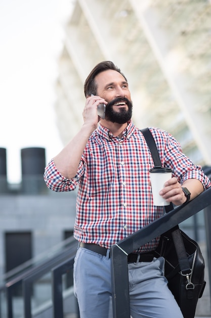 Full of joy. Cheerful pleasant man standing outdoors while having a conversation on phone