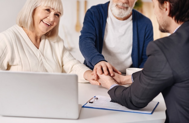 Pieno di gioia. sorridendo felice coppia di anziani seduti a casa e concludendo un accordo con l'agente immobiliare mentre si stringono la mano ed esprimono felicità