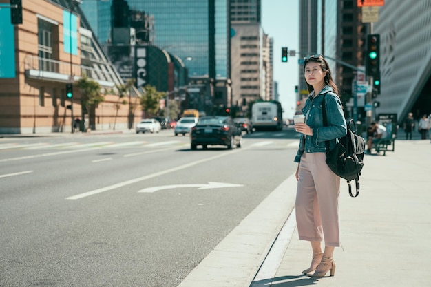 タクシーを探しているコーヒーカップを保持している道路に立っているバックパックとエレガントな服装でフルレングスのアジアの実業家。自信を持って女性起業家がオフィスに急いで大都市でイエローキャブを待っています