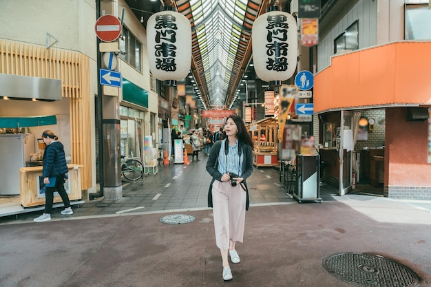 full length of asian woman photographer standing at the exit of kuromon ichiba market and looking around in Osaka japan. translation on lantern:â kuromon ichiba marketâ