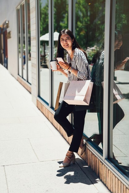 Photo full length of asian woman spending leisure time in shopping mall in stanford. young girl standing relying against by fashion store window display cloth shop. female holding coffee cellphone bags.