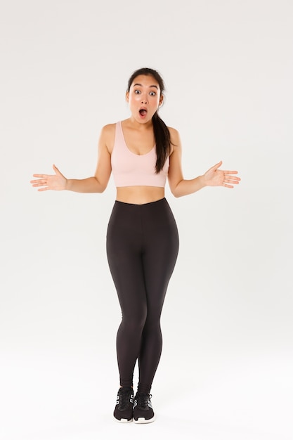 Full length of astonished and surprised female athlete, sportswoman in activewear looking impressed, drop jaw and spread hands sideways see great gym discount, workout equipment in sale.