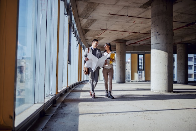 Full length of attractive caucasian architects walking in future business center and talking about fresh ideas for building. Man holding blueprints while woman holding paperwork.