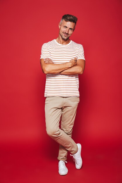 Photo full length of attractive man in striped t-shirt smiling and standing with arms crossed isolated on red