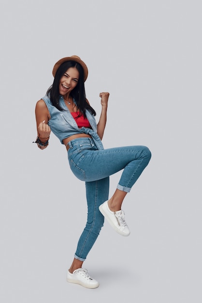 Full length of attractive stylish young woman cheering and smiling while standing against grey background