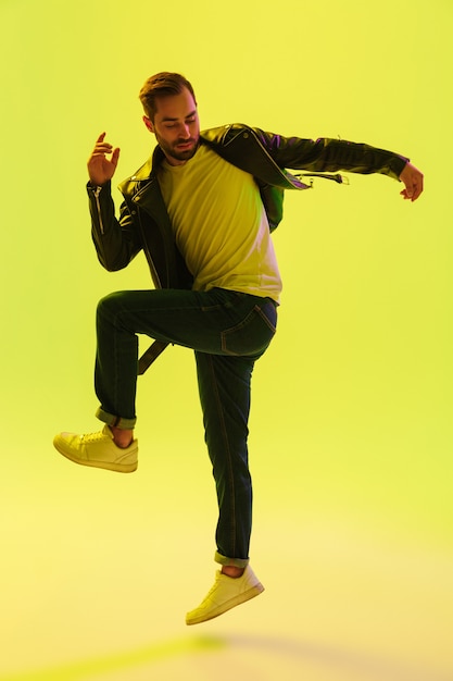 Full length of an attractive young man wearing leather jacket jumping isolated over yellow wall