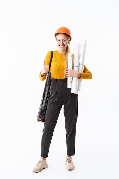 Full length of an attractive young woman architect standing isolated over white wall, carrying tube, holding house scheme, giving thumbs up