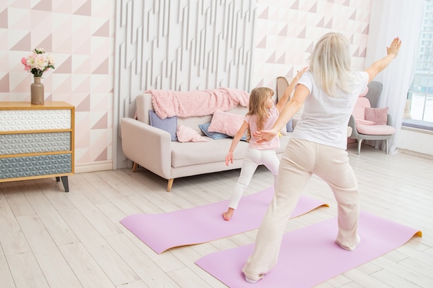 Full length back side view smiling blond mother on yoga mat with cute playful little preschool daughter do various exercises.