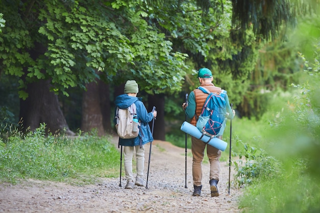 Full length back view portrait of father and son hiking together walking on wild path and carrying backpacks, copy space