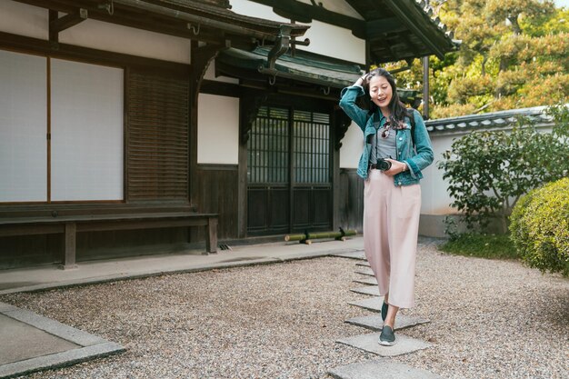 Full length of backpacker lens man walking on stone rock road
in front ground of valley of the temples. young girl traveler
holding camera cheerfully standing near japanese traditional wooden
house