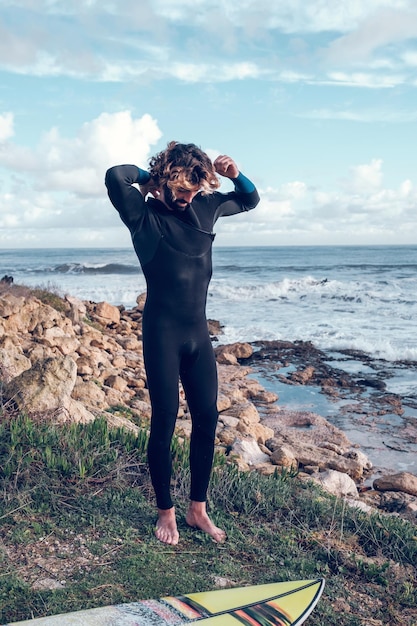 Full length of barefooted athletic Hispanic guy standing on rocky cliff near board while preparing for surfing in wavy ocean