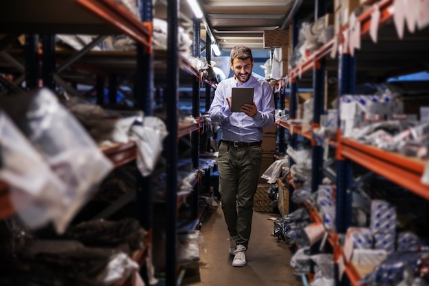 Photo full length of bearded supervisor walking trough storage of export firm, checking on goods and using tablet.