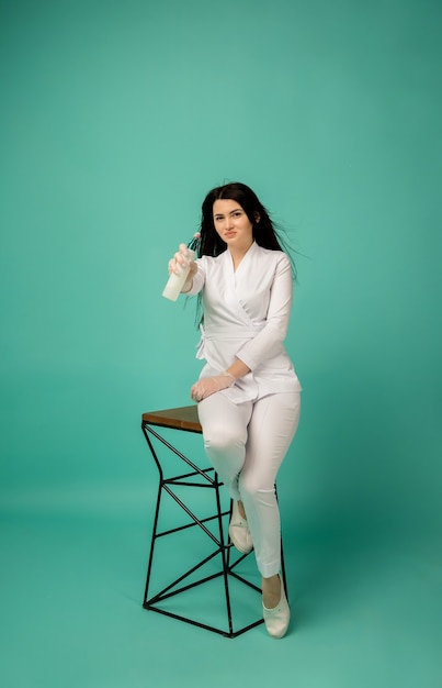 Full-length beautician in a white uniform sits on chair with a bottle on blue 