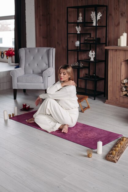 Full length of beautiful calm woman in white embracing herself sitting on pink yoga mat barefoot and looking down. Comfortable wooden interior.