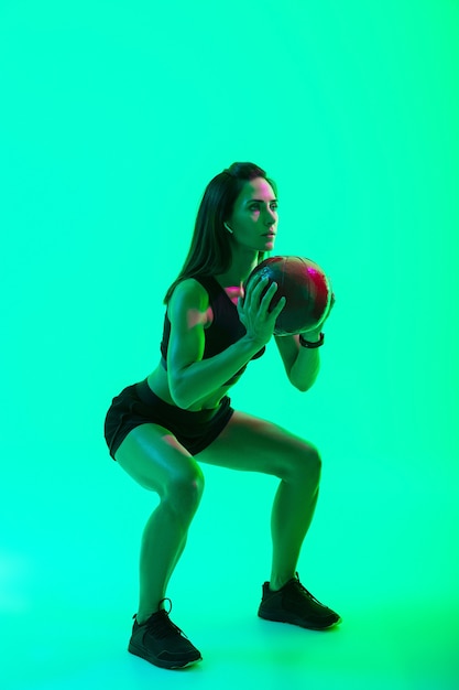 Full length of a beautiful confident young fitness girl standing isolated over green neon wall, wearing wireless earphones, working out with a medicine ball, squatting