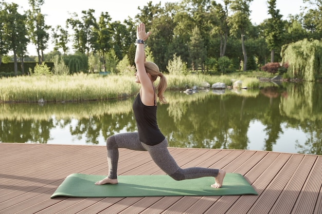 Полная длина красивой фитнес-женщины в спортивной одежде, практикующей йогу на коврике возле озера на солнечной стороне