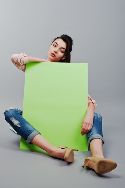 Full length of beautiful girl sitting, holding green blank advertising board banner, over gray background