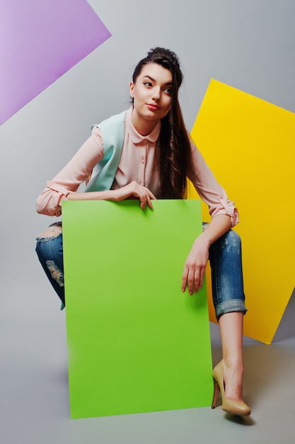 Полная длина красивая девушка сидит, держа зеленый пустой рекламный щит, на сером фоне и желтый и фиолетовый баннер
