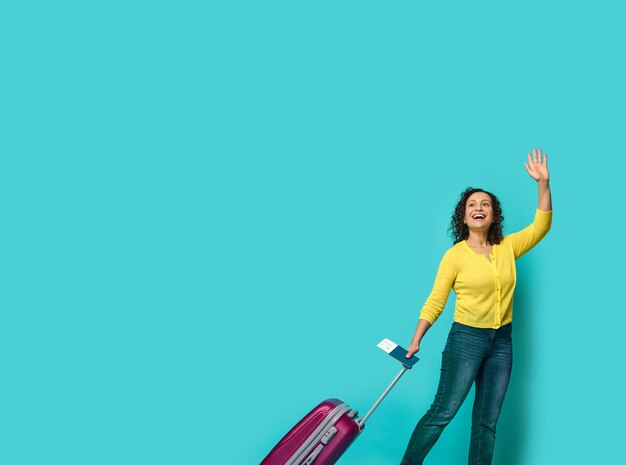 Full length of beautiful young woman in casual clothes, waving hand, holding identity document and boarding pass, posing with suitcase, smiling looking to the side at a blue background with copy space