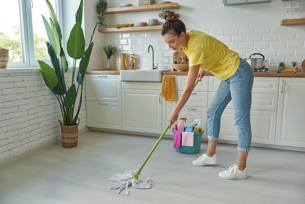 Photo full length of beautiful young woman cleaning floor with mop while standing at the kitchen