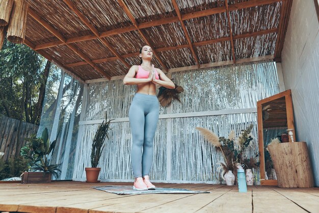 full length of beautiful young woman in sports clothing meditating on patio