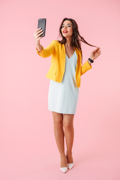 Full length of a beautiful young woman wearing colorful clothes standing isolated over pink, taking a selfie