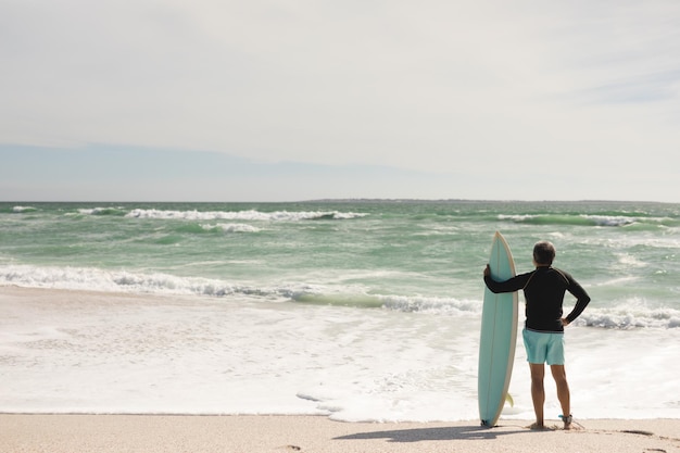 Full length of biracial senior man holding surfboard standing on shore at beach during sunny day