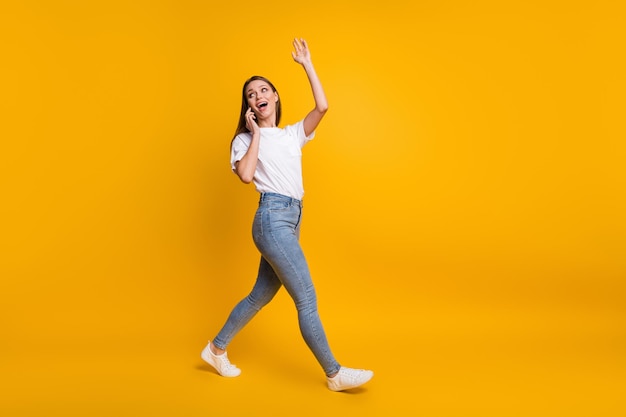 Full length body size photo of girl talking on mobile phone waving with hand greeting isolated on vivid yellow color background