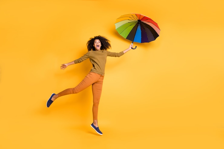 Photo full length body size photo of wavy cheerful cute nice charming pretty girlfriend flying with umbrella wearing orange pants trousers footwear isolated over vivid yellow color background