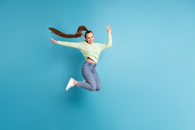 Фотография сбоку в полный рост прыгающей девушки с длинными волосами, показывающая V-образный знак на ярком синем фоне